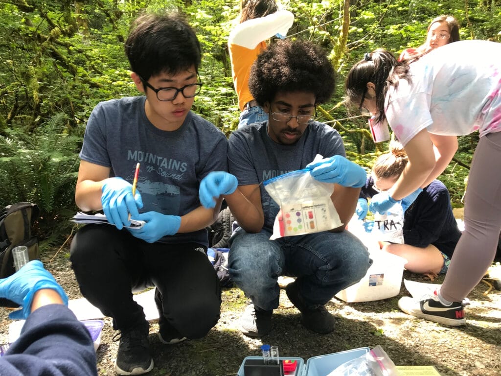 Two students crouching side-by-side and looking at a kit for science testing. One is holding a notebook and pencil.