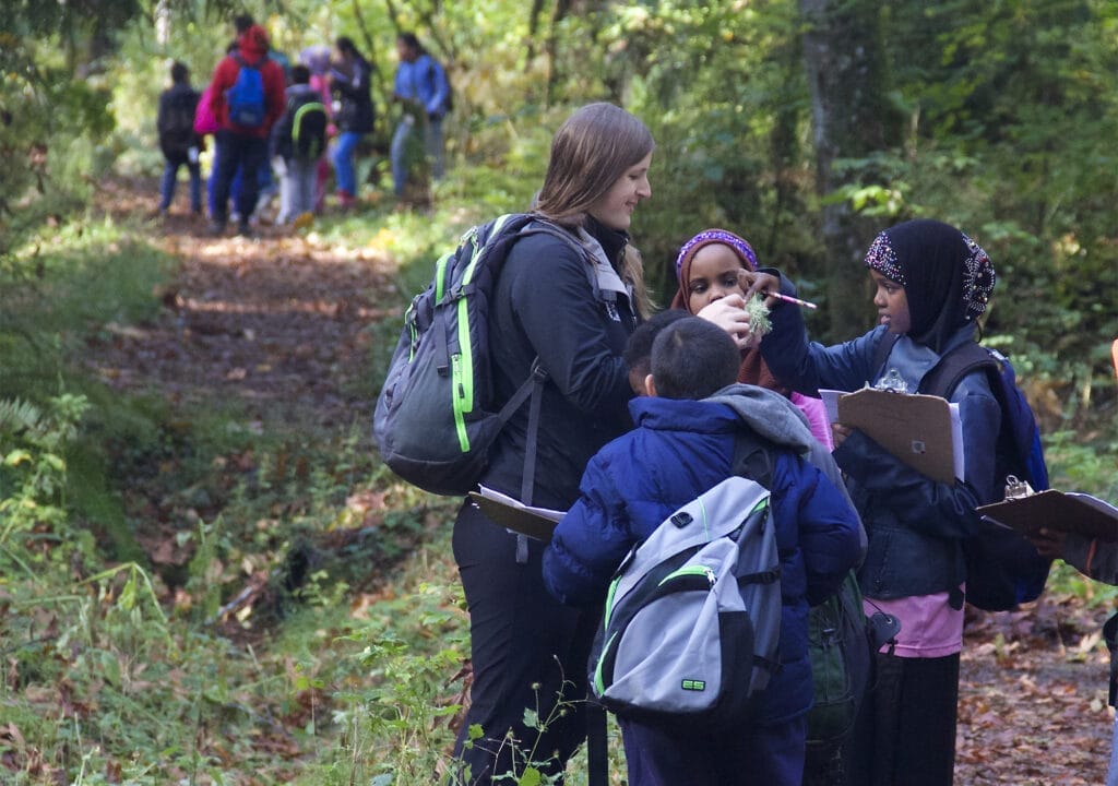 A group of students out in nature looking at something with a teacher as part of an environmental education program field trip. There is a second group of students in the background.