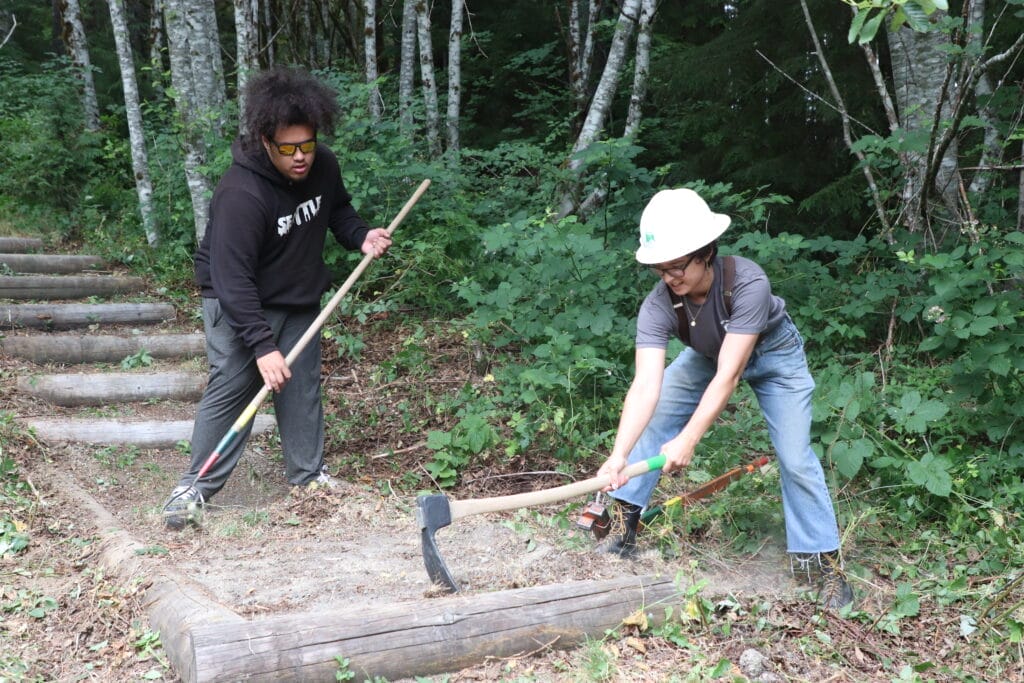 Two young adults using tools to remove weeds from an outdoor staircase along a trail.