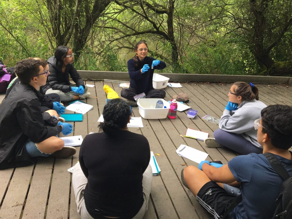 A group of students sitting on a board walk in a circle, looking at a teacher who is showing them something in a science tube, wearing gloves.