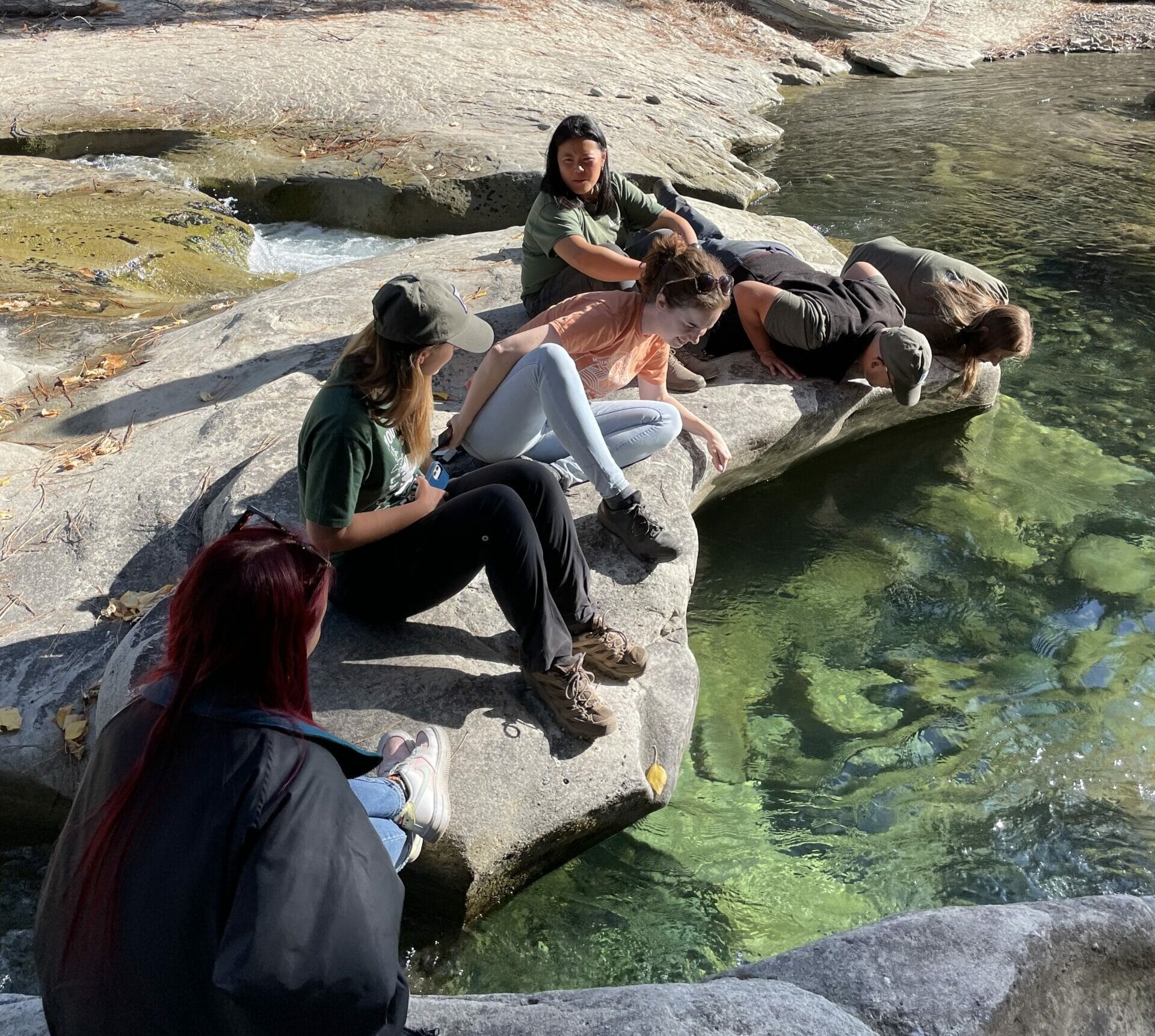 A group of six people sit on the sandstone bank of a clear river.
