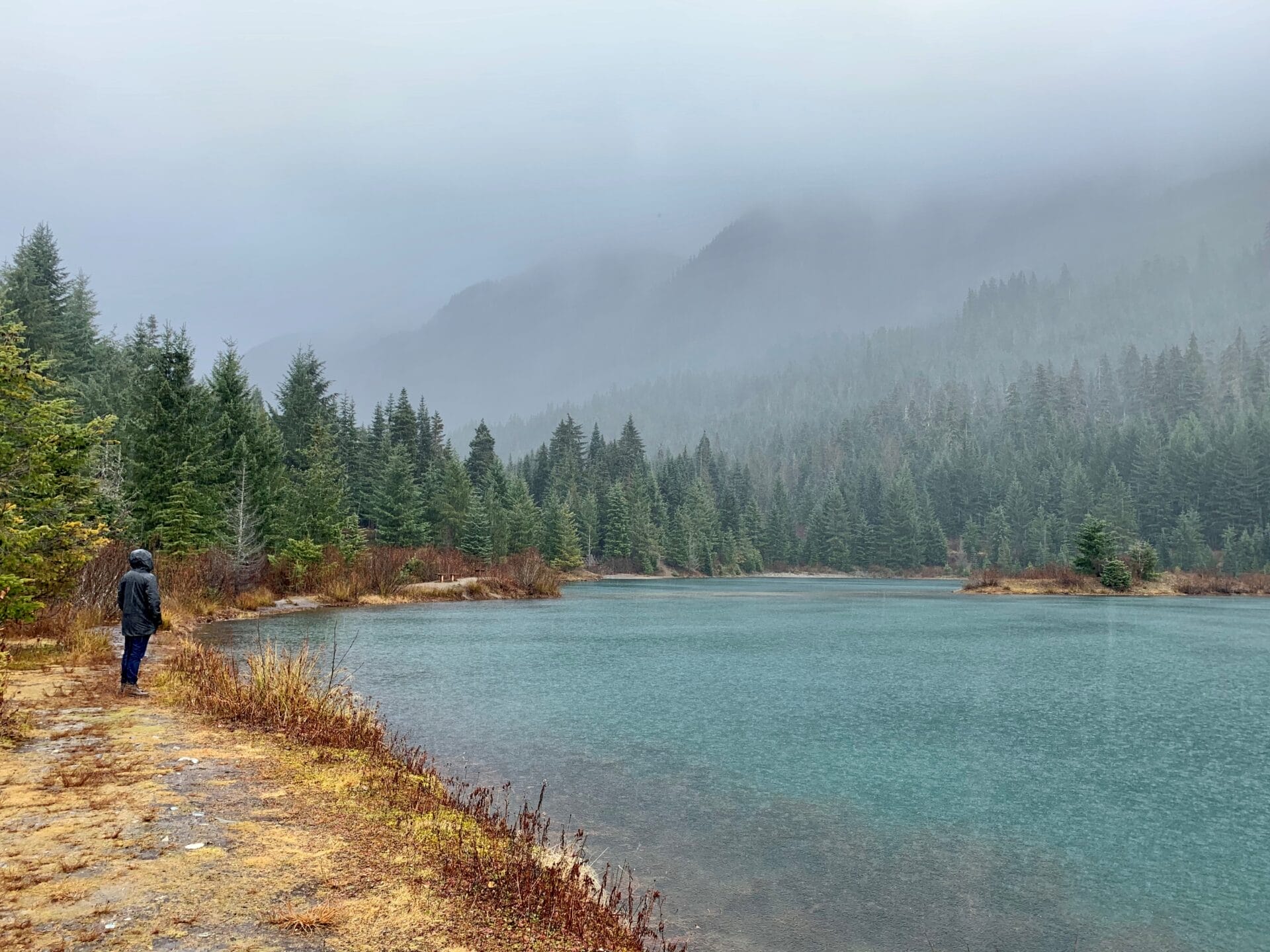 A person walks along a large turquoise pond in a misty mountainous area.